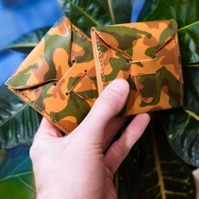 A hand holding two camouflage-dyed leather pick cases in front of a houseplant